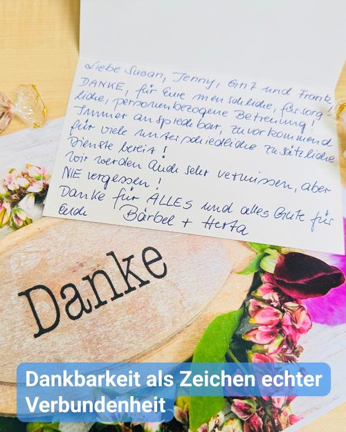 Eine Nahaufnahme zeigt eine emotionale Ausdrucksform der Dankbarkeit, präsentiert auf einer hölzernen Tafel mit dem hervorgehobenen Wort „Danke“. Die Tafel liegt eingebettet in zarte Blumengestecke, die dem Arrangement eine persönliche Eleganz und behagliche Wärme verleihen.  Eine handgeschriebene Karte, scheinbar ein Dankesbrief, überdeckt teilweise den oberen Bereich der Holztafel. Die in blauer Tinte verfasste Botschaft richtet sich an „Susan, Jenny, Gnt und Frank“ und drückt aufrichtig ihre Wertschätzung für deren fürsorgliche und aufmerksame Unterstützung aus. Die Handschrift strahlt Ehrlichkeit und Dankbarkeit aus und unterstreicht den positiven Einfluss, den die Empfänger auf die Verfasser ausgeübt haben.  Am unteren Bildrand kündigt ein blaues Banner den Satz „Dankbarkeit als Zeichen echter Verbundenheit“ an, was das zentrale Leitmotiv der Szene unterstreicht.