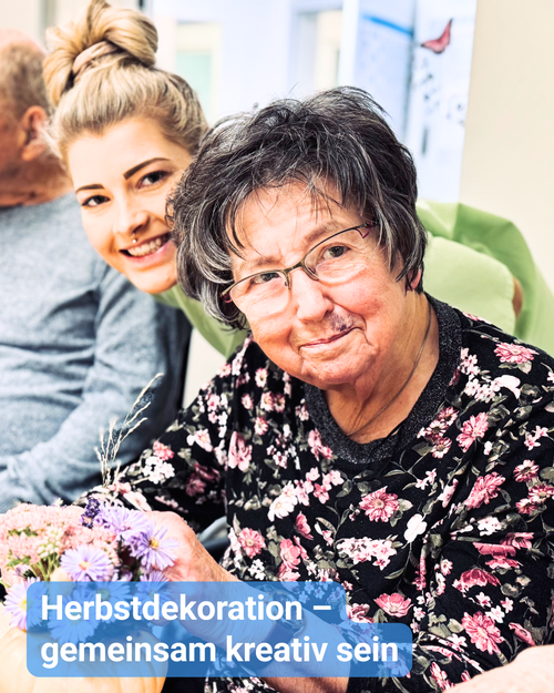 An einem großen Tisch mit Herbstdekoration sitzen ältere Menschen, und zwischen ihnen lächelt eine Betreuungskraft in die Kamera. Bildbeschriftung: Herbstdekoration – gemeinsam kreativ sein
