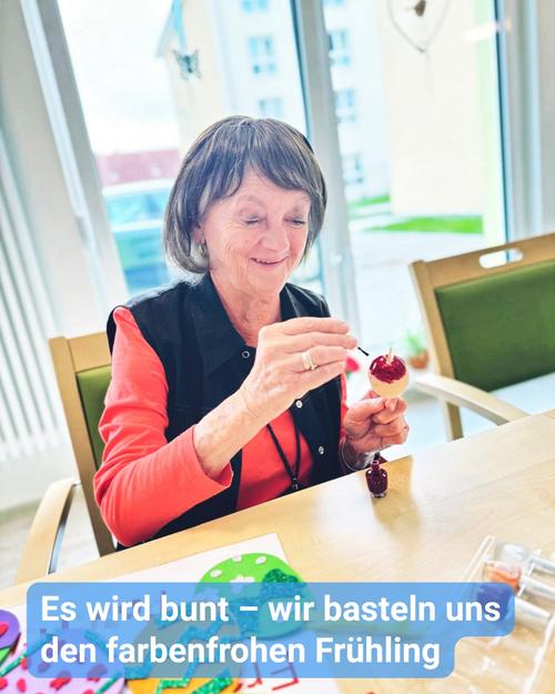 Nahaufnahme einer älteren Frau mit kurz geschnittenem, graubraunem Haar, die an einem Tisch sitzt und sich einer Bastelaktivität widmet. Sie trägt ein korallenfarbenes, kurzärmliges Shirt und eine dunkle Weste. Sorgfältig verwendet sie ein kleines Werkzeug, um eine kleines, rot gefärbtes Osterei zu dekorieren. Der Tisch besticht durch helles Holz, und verschiedene Bastelmaterialien sind sichtbar, darunter eine kleine Dose Bastelkleber sowie farbenfrohe Bastelutensilien, die auf die Kreation frühlingshafter Dekorationen hinweisen. Im Hintergrund erhellt ein strahlendes Fensterlicht den Raum und vermittelt eine fröhliche, möglicherweise häusliche Atmosphäre.  Der Hintergrund ist verschwommen, zeigt jedoch einen sonnendurchfluteten Raum mit großen Fenstern, die einen teilweise sichtbaren Außenbereich preisgeben. Der Raum wirkt sauber und ist Teil eines Seniorenzentrums. Hinter der Frau steht teilweise ein hölzerner Stuhl.  Ein überlagert eingefügter Untertitel am unteren Bildrand lautet: "Es wird bunt – wir basteln uns den farbenfrohen Frühling."